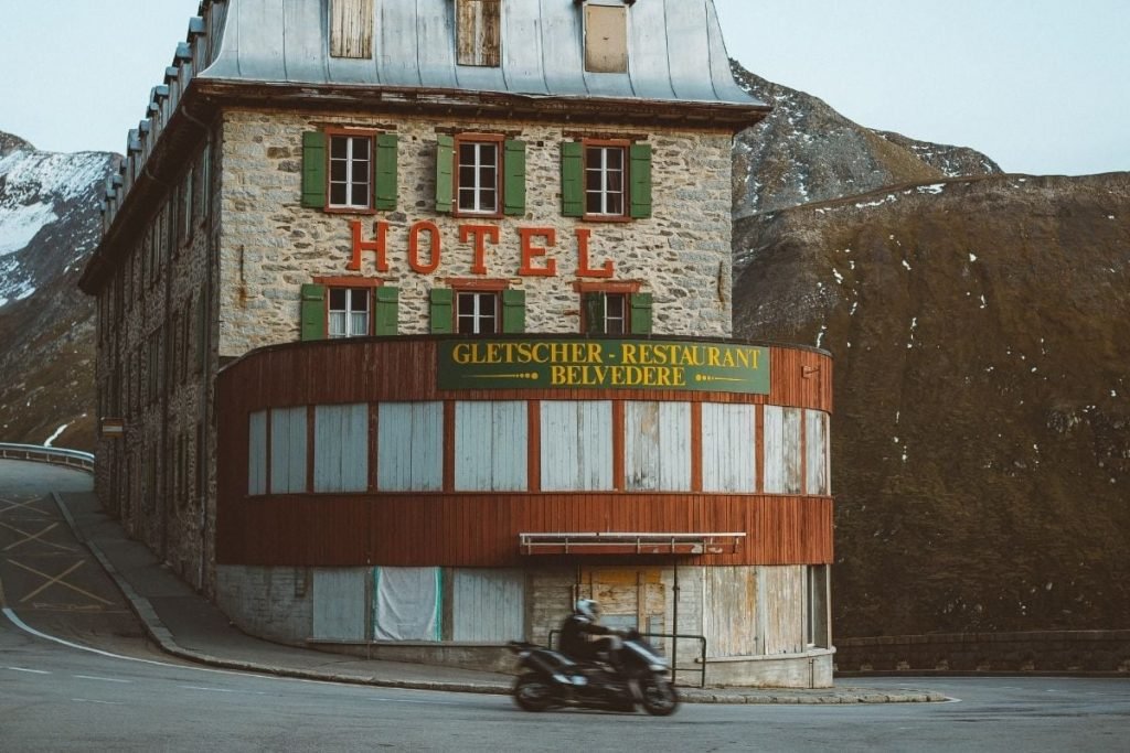 biker riding past hotel in the swiss alps - motorcycle touring on a budget